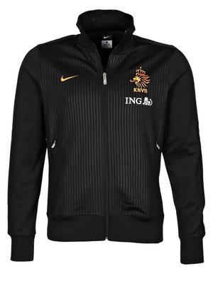 Schwarze Jacke mit durchgehendem Reißverschluss und dezenten vertikalen Streifen, mit orangefarbenem Nike-Swoosh, KNVB-Löwenemblem und weißem ING-Logo auf der Brust.