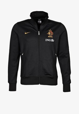 Veste noire zippée intégrale avec des rayures verticales subtiles, avec un swoosh Nike orange, un emblème de lion KNVB et un logo ING blanc sur la poitrine.