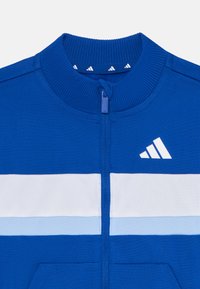 Blauwe ritsjack met geribde kraag, met witte en lichtblauwe horizontale strepen en een wit logo op de borst. Soepele textuur.