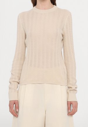 Femme portant un pull beige en maille texturée à manches longues et un pantalon large crème taille haute, debout devant un fond blanc.