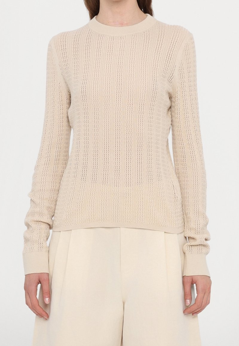 Femme portant un pull beige en maille texturée à manches longues et un pantalon large crème taille haute, debout devant un fond blanc.