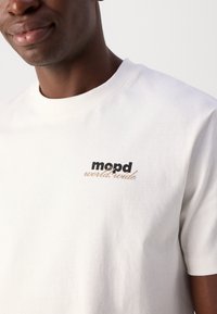 Balts kokvilnas T-krekls ar apaļu dekoltē, īsām piedurknēm un melnu un zelta teksta logo "mopd world.wide" kreisajā krūtīs.