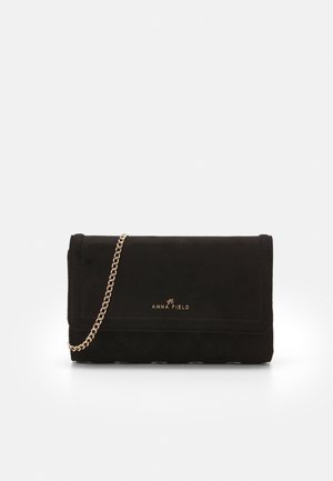 Schwarze Wildleder-Clutch mit goldener Kettenriemen und "ANNA FIELD" Logo in Gold auf der Vorderklappe vor schlichter Hintergrund.