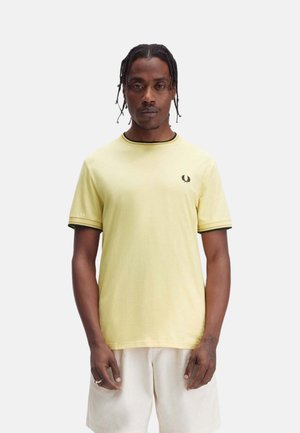 Hombre con el pelo trenzado, que lleva una camiseta de manga corta amarillo pálido con ribete negro y pantalones cortos blancos, de pie contra un fondo blanco liso.