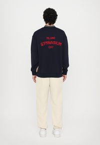 Sweatshirt azul-marinho com "MILANO GYMNASIUM EA7" bordado em vermelho nas costas, punhos canelados e corte descontraído, combinado com calças bege e sapatos brancos.