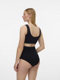 Ženska s ravnimi blond lasmi, oblečena v črn brezrokavnik in črne hlačke visoke pasu, obrnjena bočno in gleda levo.