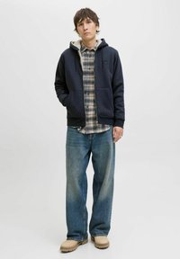 Granatowa kurtka z kapturem z przednimi kieszeniami nałożona na koszulę w kratę, zestawiona z luźnymi niebieskimi dżinsami i beżowym obuwiem. Prosty i casualowy design.