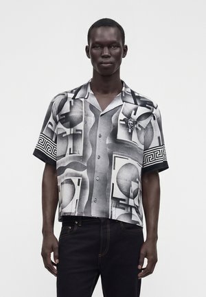 Uomo che indossa una camicia a maniche corte bianca e nera con motivi geometrici e una spilla a forma di testa di Medusa sulla tasca del petto.
