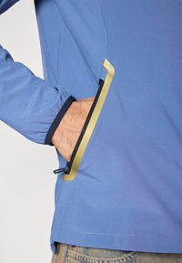 Μπλε μπουφάν με τσέπη που κλείνει με φερμουάρ, τονισμένη με κίτρινη λεπτομέρεια. Διαθέτει ελαστικά μανίκια και απαλή, ελαφριά υφή.