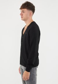 Cardigan nero con scollo a V, bottoni frontali e silhouette aderente. Realizzato in tessuto morbido con maniche lunghe, dallo stile minimalista.