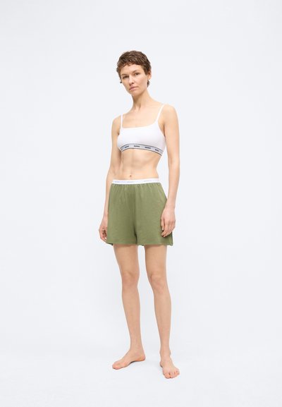Femme debout pieds nus portant un débardeur court blanc et un short ample vert olive sur un fond clair uni.