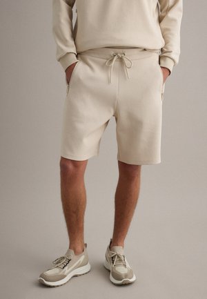 Hombre con pantalones cortos beige con cordón, sudadera beige y zapatillas beige, de pie con las manos en los bolsillos contra un fondo liso.