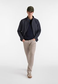 Chaqueta navy, suéter oscuro, pantalones beige y zapatillas marrones. El modelo se encuentra de manera casual, con las manos en los bolsillos y usando una gorra verde.