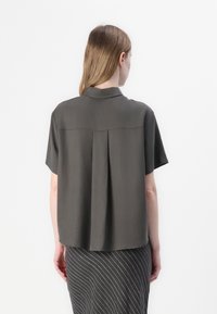 Camicia a maniche corte con colletto in tessuto grigio scuro. Presenta una piega sul retro, design leggermente accorciato e texture liscia.