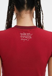 Rød tætsiddende t-shirt med rund hals og korte ærmer, med hvid tekst "Don Ed Hardy designs" trykt på bagsiden.