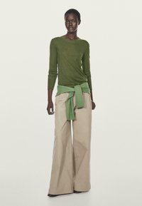 Maglietta verde a maniche lunghe sovrapposta a un maglione verde, abbinata a pantaloni beige larghi. Le trame morbide e gli elementi di design semplici sono in evidenza.