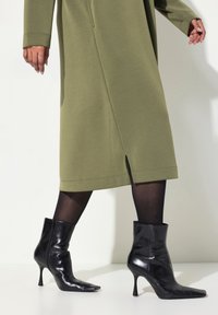 Abito verde oliva con vestibilità morbida e spacco laterale, abbinato a stivaletti neri con punta a punta e tacco a stiletto con finitura lucida.