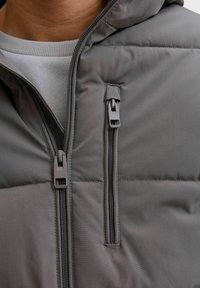 Zbliżenie osoby w szarej, pikowanej kurtce na jasnoszarym swetrze, z dwoma pionowymi kieszeniami na zamek na kurtce.