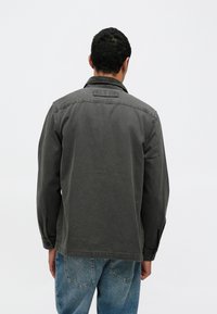 Γκρι βαμβακερό μπουφάν με μακριά μανίκια και διπλωμένο γιακά. Στο πίσω μέρος υπάρχει κεντημένο λογότυπο και μανσέτες με κουμπιά. Φοριέται με ανοιχτόχρωμο μπλε τζιν.