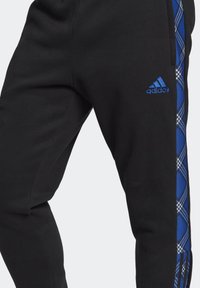 Pantaloni sportivi neri con pannelli laterali a quadri blu e logo Adidas blu ricamato sul davanti. Tessuto morbido con elastico in vita.