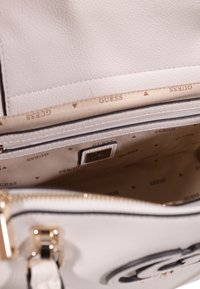 Borsa a tracolla in pelle bianca con superficie texture, dotata di una tasca con zip e un interno beige rivestito con la parola "GUESS" in un motivo ripetuto.
