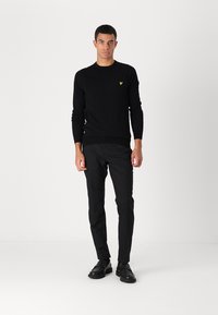 Un pull noir en tricot avec un col rond et des poignets côtelés, featuring un petit logo jaune sur la poitrine gauche, associé à un pantalon noir et des chaussures.