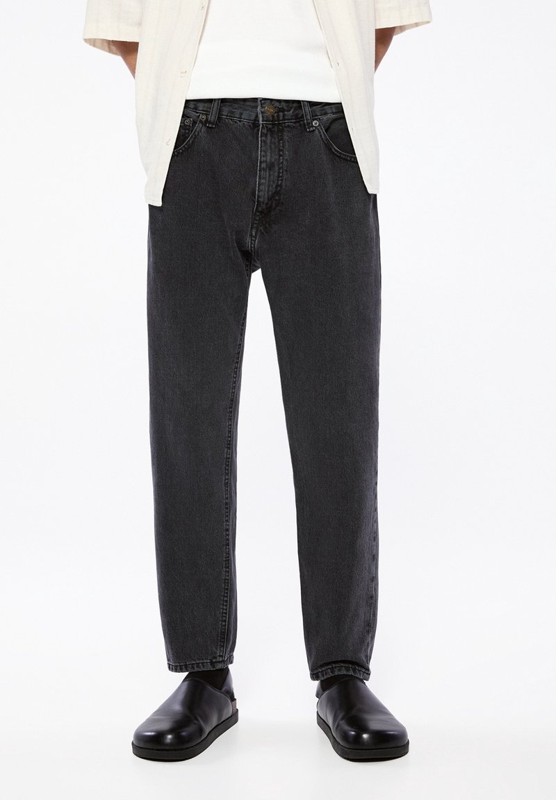 Person trägt schwarze Jeans mit lockerem Sitz, schwarze Slipper und ein cremefarbenes offenes Hemd über einem weißen Oberteil, steht vor weißem Hintergrund.