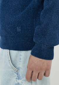 Pull en tricot bleu marine avec des poignets côtelés, arborant un petit logo sur l'ourlet. La texture semble moelleuse et douce. Porté sur un jean en denim clair.