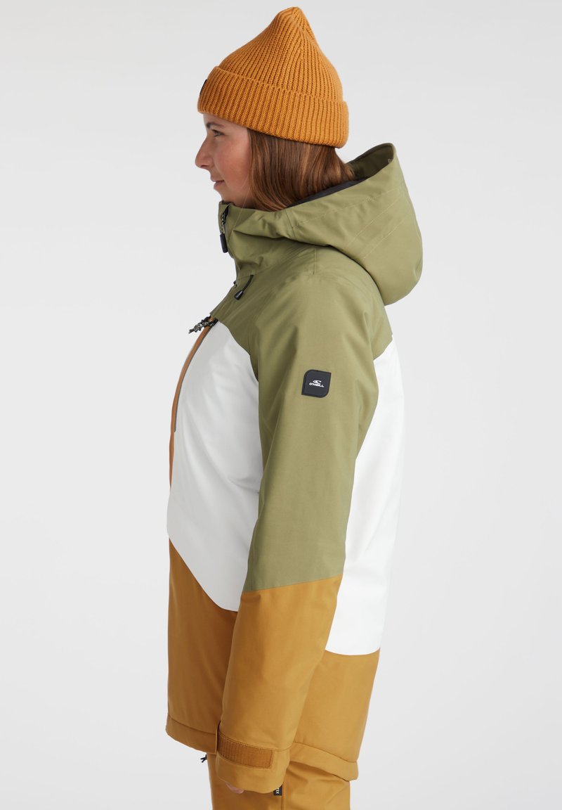 Frau in einer dreifarbigen Winterjacke und einer senfgelben Strickmütze, im Profil nach links vor einem schlichten Hintergrund.