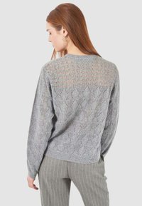 Donna con lunghi capelli rossi che indossa un maglione lavorato a maglia grigio chiaro con un motivo e pantaloni grigi a righe, rivolta verso un sfondo neutro.