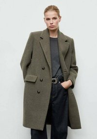 Abrigo de lana verde oliva con doble botonadura, grandes botones, solapas de chal y bolsillos laterales, llevado sobre una blusa gris y vaqueros oscuros.