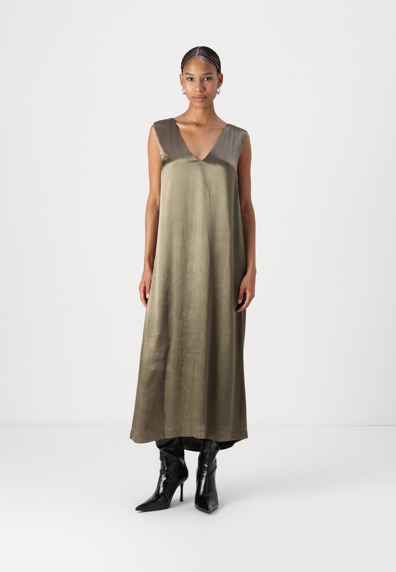 Robe en satin olive vert longue avec un décolleté en V profond et sans manches. Associée à des bottes noires à talons pointus montant jusqu'au genou. Texture lisse.