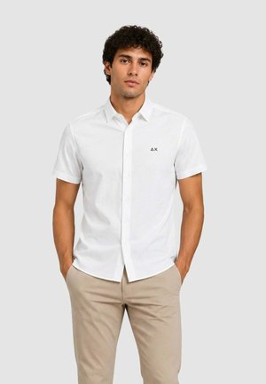 Uomo con capelli ricci che indossa una camicia bianca a maniche corte con un piccolo logo sul petto e pantaloni beige, mani in tasca.