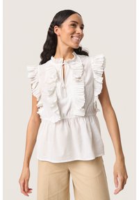 Hvid ærmeløs bluse med flæset blondekant langs udskæringen og skuldrene. Elastisk talje og blødt stof, parret med beige bukser.
