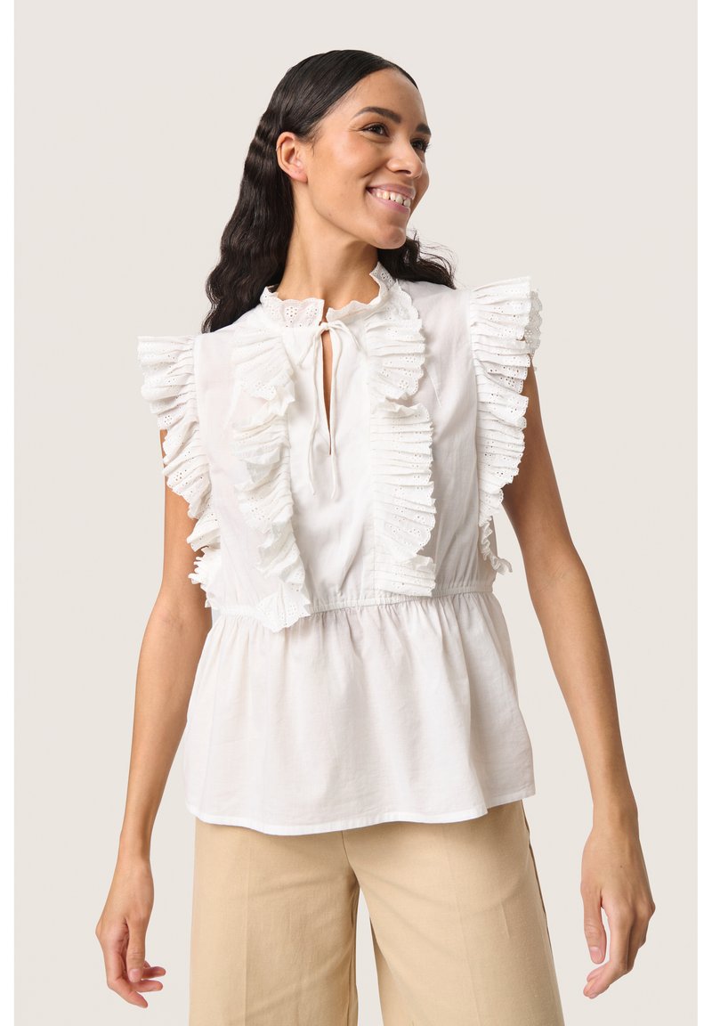 Hvid ærmeløs bluse med flæset blondekant langs udskæringen og skuldrene. Elastisk talje og blødt stof, parret med beige bukser.