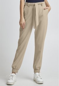 Beige, schmal zulaufende Hose mit gebundenem Bund, seitlichen Taschen und gerippten Bündchen an den Knöcheln. Kombiniert mit weißen Sneakern für einen Kontrast.