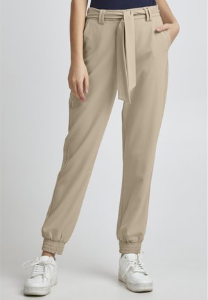 Pantaloni beige a sigaretta con vita legata, dotati di tasche laterali e polsini a coste. Abbinati a sneaker bianche per un contrasto.