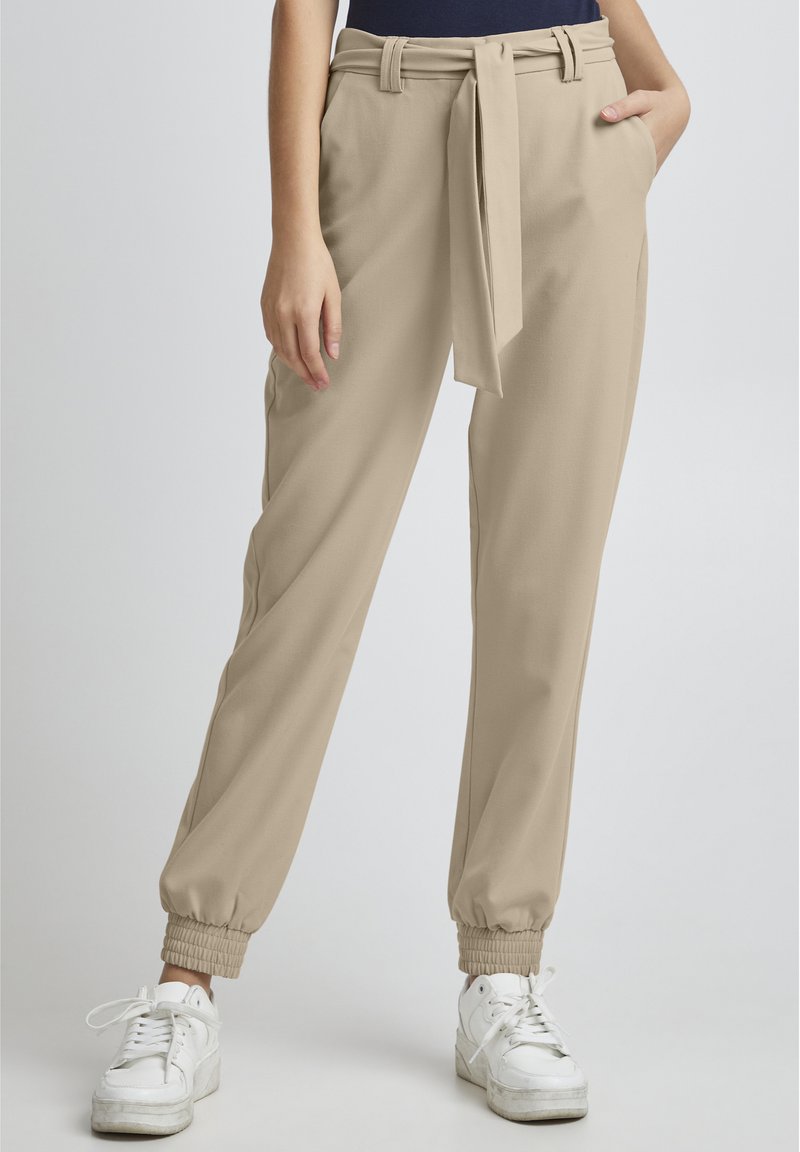 Beige, schmal zulaufende Hose mit gebundenem Bund, seitlichen Taschen und gerippten Bündchen an den Knöcheln. Kombiniert mit weißen Sneakern für einen Kontrast.