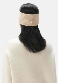 Beige ribbat stickad pannband tillverkat av mjukt material med en liten etikett, bärs på mörkt hår och täcker öronen.