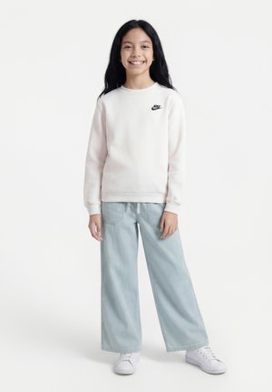 Lächelndes Mädchen mit langen dunklen Haaren trägt weißen Nike-Sweatshirt, hellblaue Jeans mit weitem Bein und weiße Turnschuhe, steht vor weißem Hintergrund.