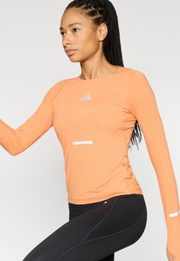 Ilgas rankovėmis oranžinis marškinėlis su apvalia iškirpte ir aptemptu kirpimu. Viršutinėje priekinėje dalyje ir apatinėje margintų atspindinčių elementų, derinamų su juodomis tamprėmis.