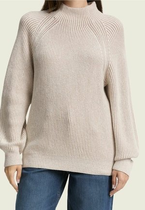 Beige ribstrikket sweater med ballonærmer og høj hals båret med blå jeans på en person.