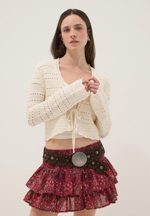 Jonge vrouw die een crèmekleurige gehaakte cardigan draagt en een rode, gelaagde minirok met patroon, gecombineerd met een brede, met studs versierde zwarte riem.