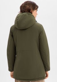 Veste matelassée vert olive avec capuche, texture lisse et détails de couture. Présente une silhouette ajustée et des éléments réglables.