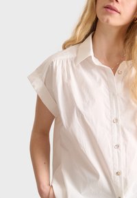Weißes Baumwollhemd mit kurzen hochgerollten Ärmeln,Knopfleiste, weichem Textil und dezenten Falten an den Schultern. Verfügt über klare Knöpfe.