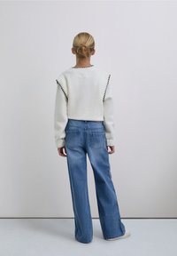 Suéter blanco con ribete negro, combinado con jeans azules de pierna ancha que presentan costuras en forma de estrella a lo largo de los laterales. Fondo neutro.