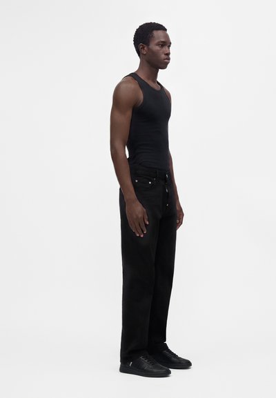 Homme debout de profil portant un débardeur noir moulant, un pantalon ample noir et des baskets noires sur un fond blanc uni.