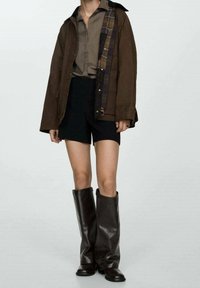 Person iført brun jakke med plaidfor, taupe skjorte, sorte shorts og høje mørke læderstøvler, der står foran en ensfarvet baggrund.