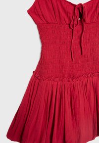 Robe rouge sans manches avec corsage smocké, col à nouer et jupe froncée avec volants.