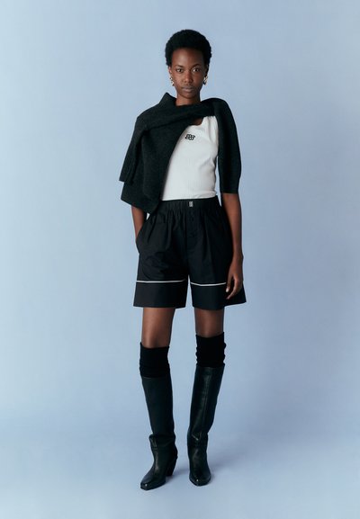 Shorts noirs avec des bordures blanches, portés avec un débardeur blanc ajusté et un pull gris foncé drapé sur les épaules. Bottes noires jusqu'aux genoux.